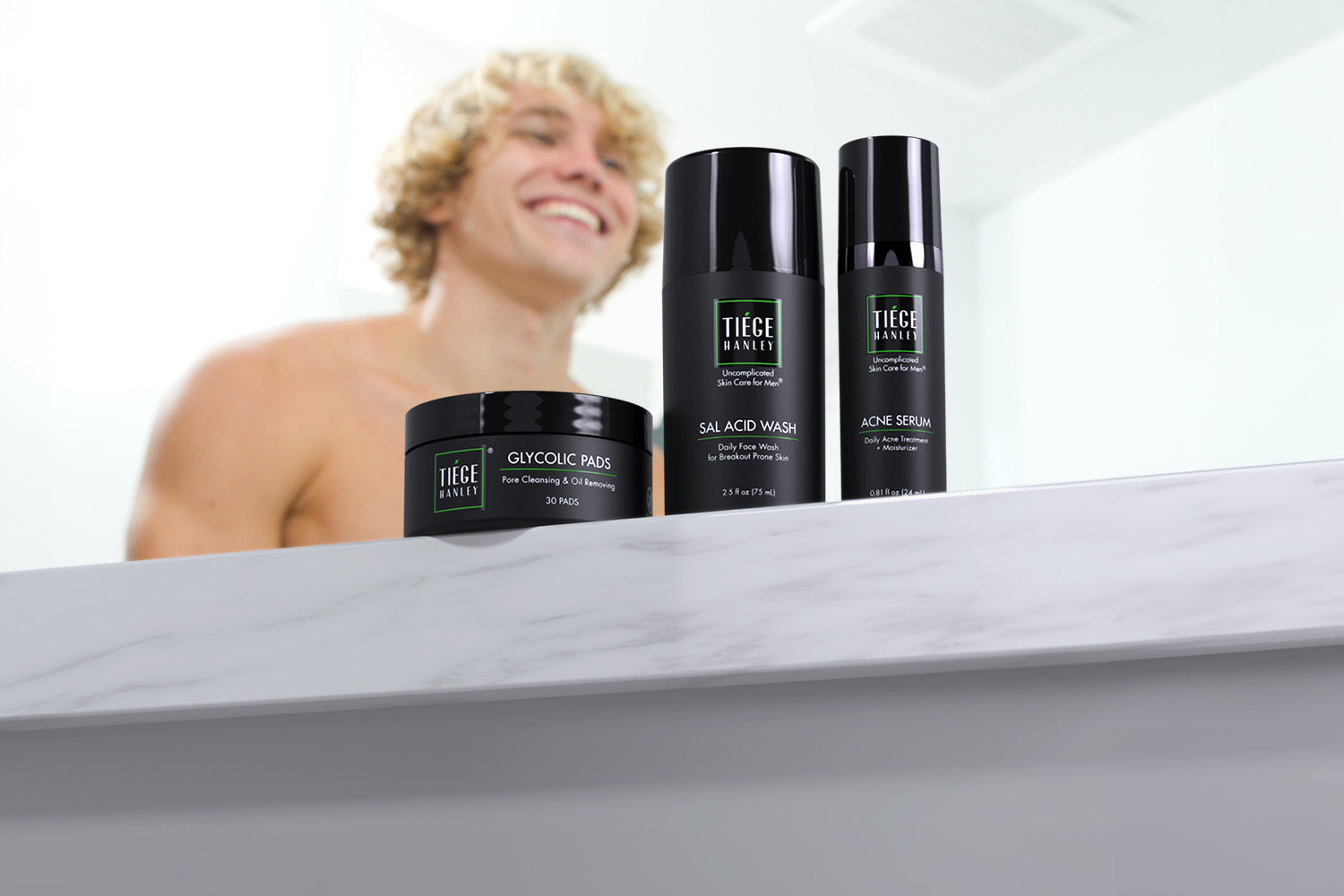 A happy man with curly blonde hair smiles at three Tiege Hanley men's skin care products, including Glycolic Pads, Sal Acid Wash, and Acne Serum, arranged on a counter.