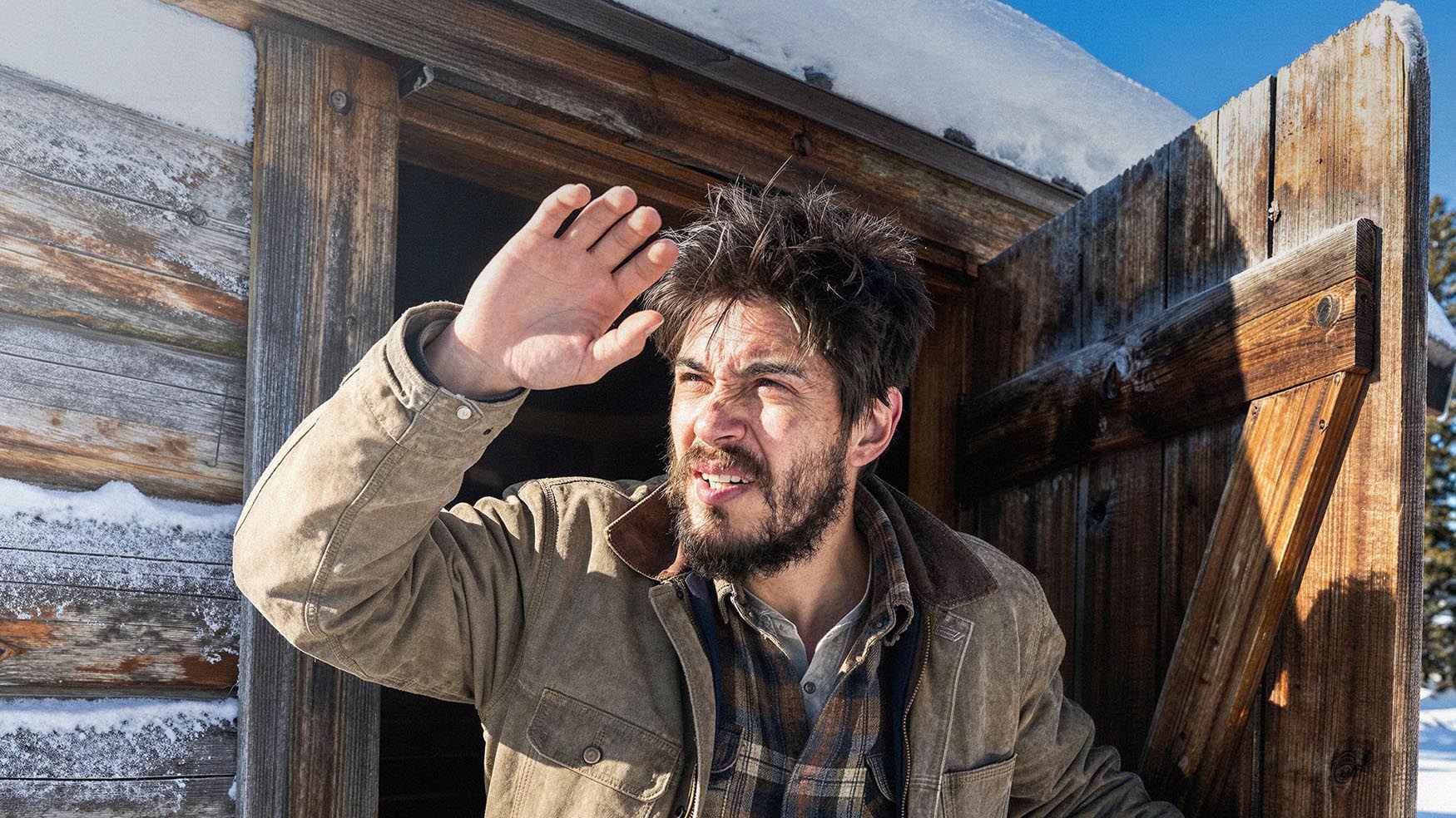 Man with a beard and mustache standing in front of a wooden cabin in a snowy landscape.