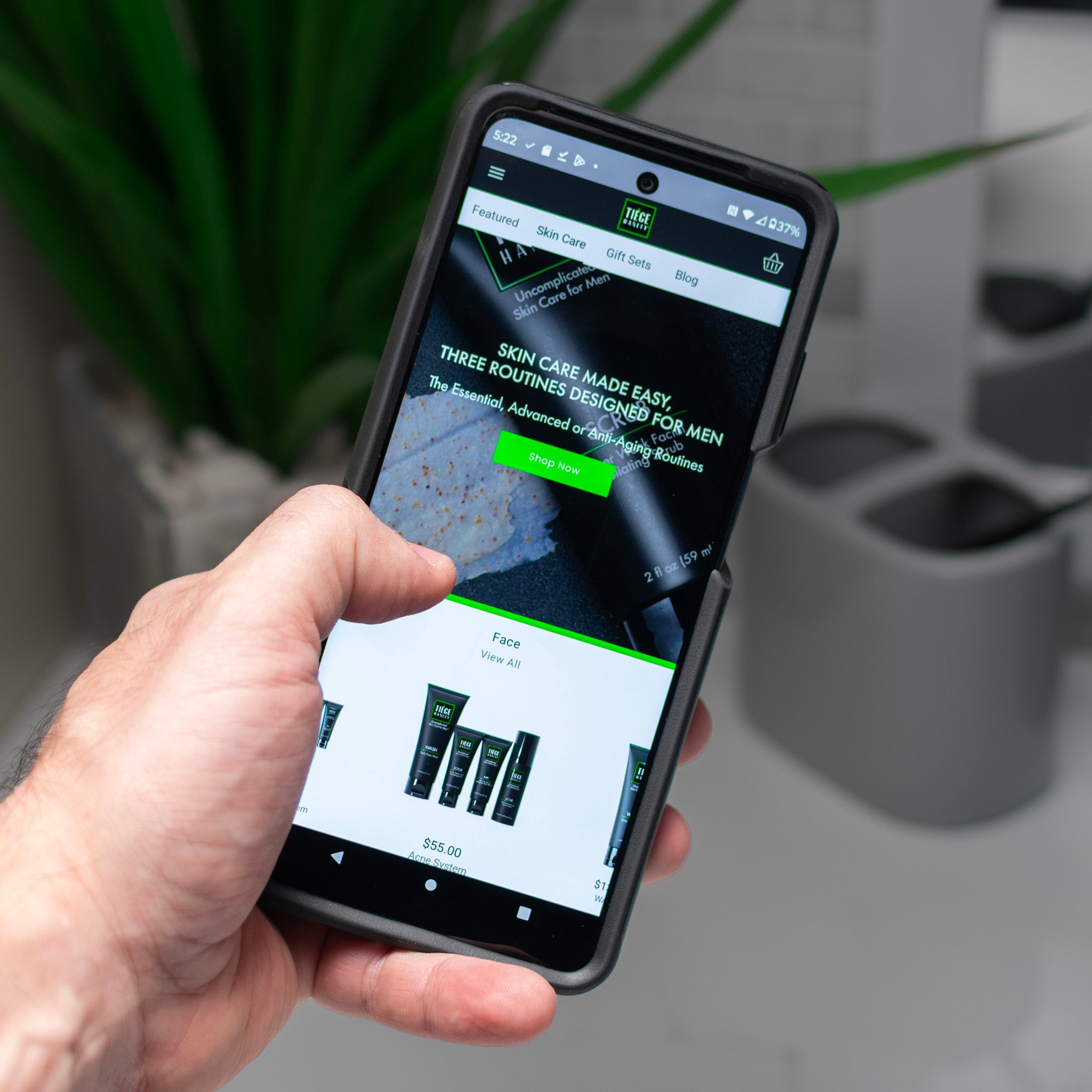 A hand holding a phone with the Tiege Hanley website on the screen.